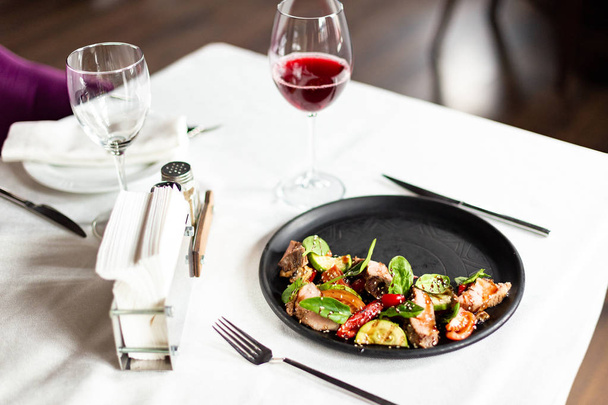 Ciepła Sałatka z cielęciny średnio palona i warzyw, podawane na czarny talerz na stole z białym obrusem, urządzeń i kieliszku wina w restauracji - Zdjęcie, obraz
