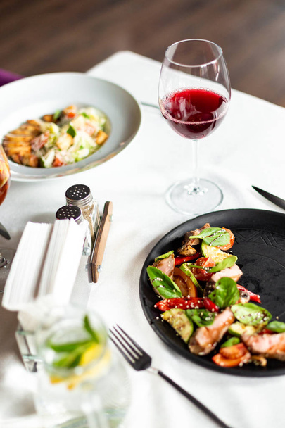 Warm salad with medium roast veal and vegetables, served on a black plate on a table with a white tablecloth, appliances and a glass of wine in a restaurant - 写真・画像