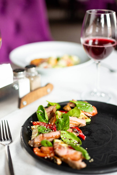 Warm salad with medium roast veal and vegetables, served on a black plate on a table with a white tablecloth, appliances and a glass of wine in a restaurant - Photo, image