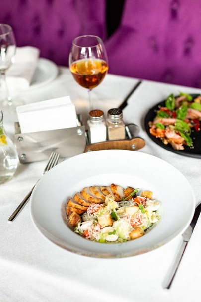 Romain salad with grilled chicken, garlic croutons and parmesan served on a white plate on a table with a white tablecloth, appliances and a glass of wine in a restaurant - Foto, afbeelding