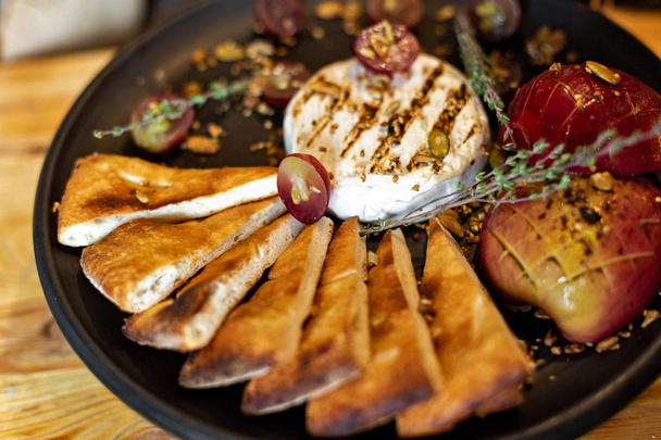Grilled camembert with baked apple and grapes with slices of bread, served on a black plate on a wooden background in rustic style. Restaurant serving - Fotoğraf, Görsel