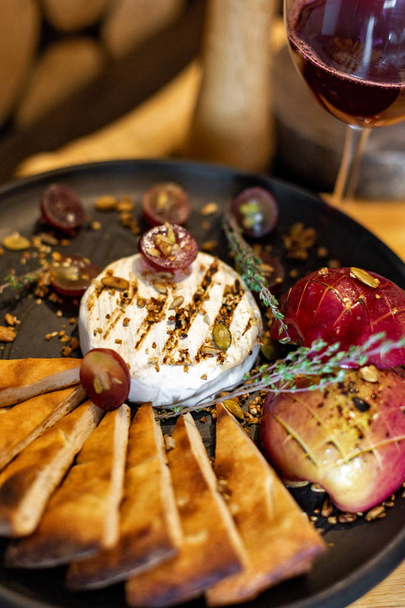 Grilled camembert with baked apple and grapes with slices of bread, served on a black plate on a wooden background in rustic style. Restaurant serving - 写真・画像