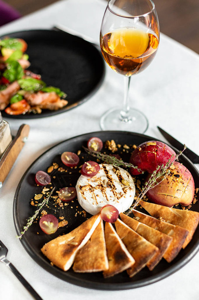 Grilled Camembert with baked apple and grapes with slices of bread tortilla, served on a black plate on a table with a white tablecloth, appliances and a glass of wine in a restaurant - Foto, afbeelding