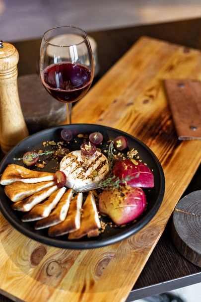 Grilled camembert with baked apple and grapes with slices of bread, served on a black plate on a wooden background in rustic style. Restaurant serving - Photo, Image