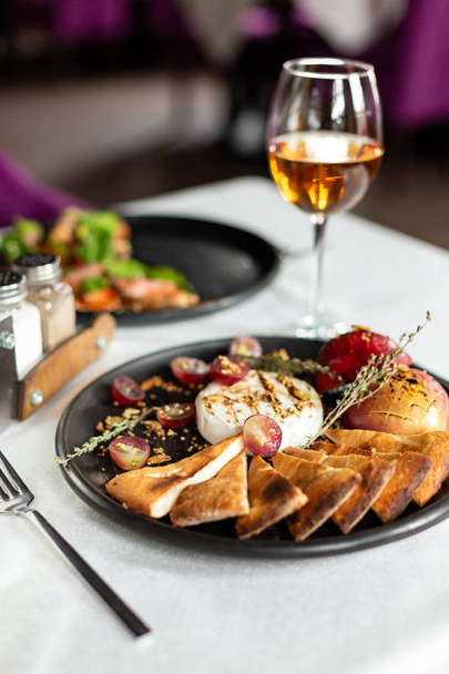 Grilled Camembert with baked apple and grapes with slices of bread tortilla, served on a black plate on a table with a white tablecloth, appliances and a glass of wine in a restaurant - Photo, image