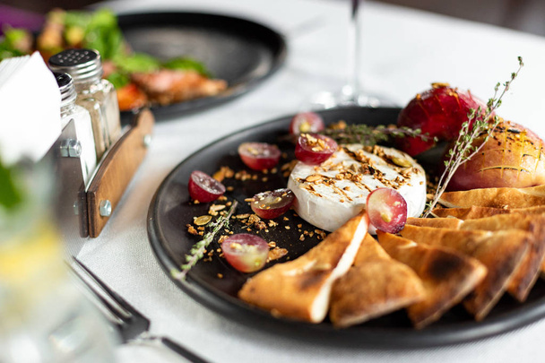 Grilled Camembert with baked apple and grapes with slices of bread tortilla, served on a black plate on a table with a white tablecloth, appliances and a glass of wine in a restaurant - Fotó, kép