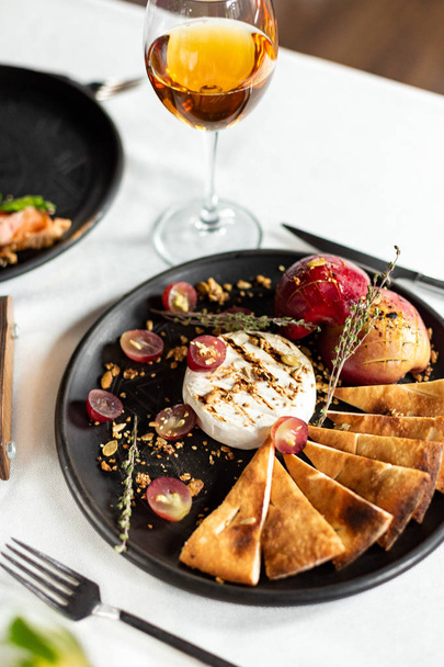 Grilled Camembert with baked apple and grapes with slices of bread tortilla, served on a black plate on a table with a white tablecloth, appliances and a glass of wine in a restaurant - 写真・画像