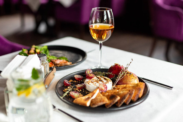 Grilled Camembert with baked apple and grapes with slices of bread tortilla, served on a black plate on a table with a white tablecloth, appliances and a glass of wine in a restaurant - Foto, Bild