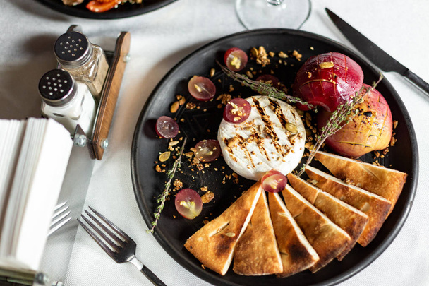 Grilled Camembert with baked apple and grapes with slices of bread tortilla, served on a black plate on a table with a white tablecloth, appliances and a glass of wine in a restaurant - Foto, Imagen