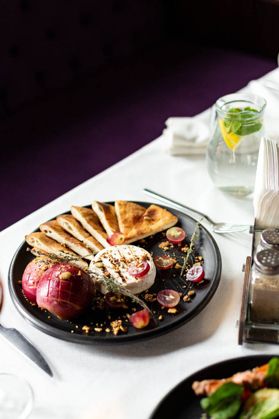 Grilled Camembert with baked apple and grapes with slices of bread tortilla, served on a black plate on a table with a white tablecloth, appliances and a glass of wine in a restaurant - Photo, Image