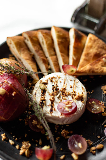 Grilled Camembert with baked apple and grapes with slices of bread tortilla, served on a black plate on a table with a white tablecloth, appliances and a glass of wine in a restaurant - 写真・画像