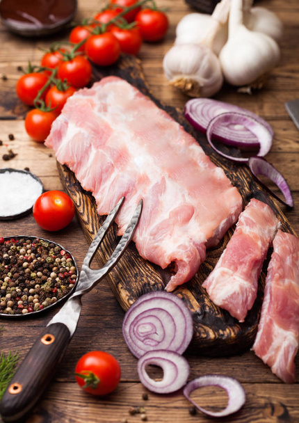 Fresh raw pieces of pork ribs on chopping board and vintage meat fork on wooden background. Fresh tomatoes and red onion with garlic salt and pepper and dill - Foto, Imagen