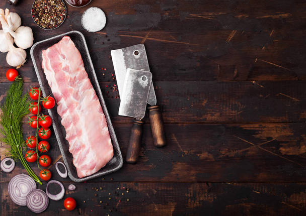 Plastic tray with raw pork ribs and vintage hatchets on wooden background. Fresh tomatoes and red onion with garlic salt and pepper.  - Fotó, kép