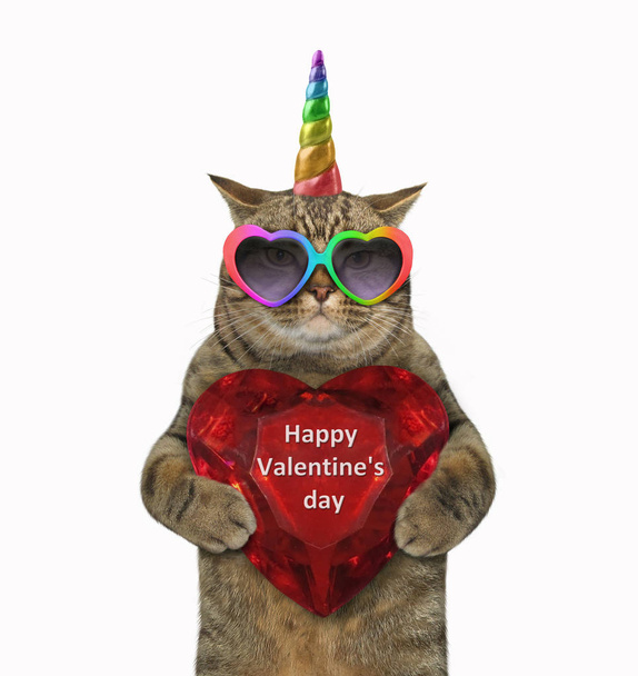 Das Katzeneinhorn mit der farbigen Sonnenbrille hält ein rubinrotes Herz mit der Aufschrift "Happy Valentinstag" in der Hand. isoliert. - Foto, Bild