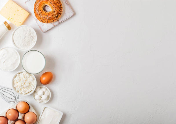 Fresh dairy products on white table background. Glass of milk, bowl of sour cream and cottage cheese and eggs. Fresh baked bagel. Steel whisk. Top view. - Foto, Imagen