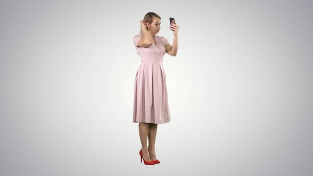 Beauty woman looking at herself in her smartphone fixing her hair on gradient background. - 写真・画像
