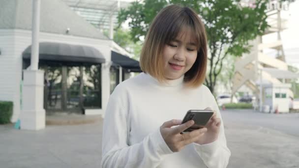 fröhliche asiatische Touristenbloggerin mit Touchscreen-Technologie am Smartphone, während sie abends auf der Straße in der Innenstadt spaziert. Lifestyle Rucksack touristisches Reisekonzept. - Filmmaterial, Video