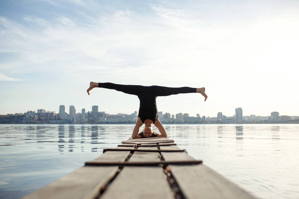 Young woman practicing yoga exercise at quiet wooden pier with city background. Sport and recreation in city rush. - Foto, Bild