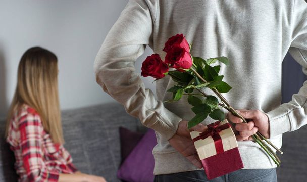 Mans hands hiding holding chic bouquet of red roses and gift with white ribbon behind back and woman with turned head awaits surprise in bed at home. Close up, selective focus - Photo, Image