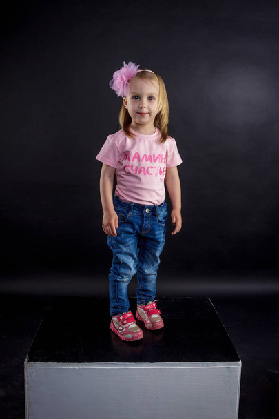 Portrait of cute European little blonde girls in jeans and pink t-shirt with the inscription: "Dad's pride, mom's joy", on a black isolated background - Foto, imagen