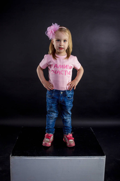 Portrait of cute European little blonde girls in jeans and pink t-shirt with the inscription: "Dad's pride, mom's joy", on a black isolated background - Foto, imagen