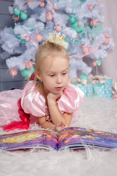 Portrait of cute european little blonde princess girl with crown in beautiful dress lying on the floor and reading book in decorated studio in christmas location with toys and christmas tree - Photo, Image