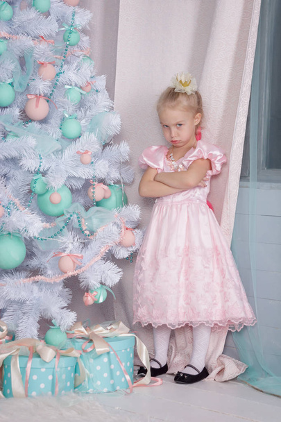 Portrait of an offended and frustrated European little blonde Princess girl with a crown in a beautiful dress and opening gifts in a decorated Studio  with toys and a Christmas tree - Photo, Image