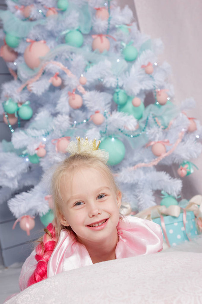 Portrait of cute european little blonde princess girl with crown in beautiful dress in decorated studio in christmas location with toys and christmas tree - Photo, Image