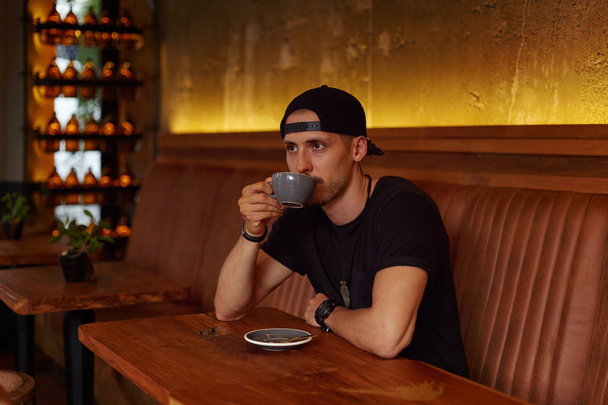 People and lifestyle concept. Attractive successful young Caucasian man drinking coffee sitting at cafe table with mug, bottles and stylish interior on background, looking through window on distance. - Photo, Image