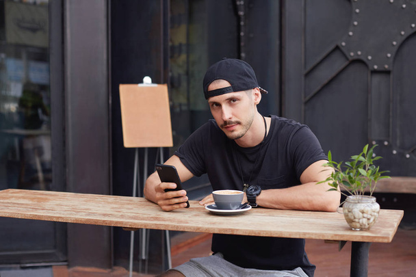 Joven hipster con sombrero negro y camiseta, bebiendo té o café y escribiendo un mensaje en un teléfono inteligente genérico. Hombre atractivo pasar un buen tiempo en la cafetería solo con wi-fi en gadget electrónico
.  - Foto, Imagen