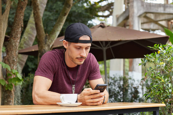 Guapo joven turista masculino americano con sombrero, accesorios de moda sonriendo suavemente mientras lee sms en el teléfono inteligente, mensajería a su novia en línea usando wi-fi gratis, tomando café en la cafetería al aire libre
. - Foto, imagen