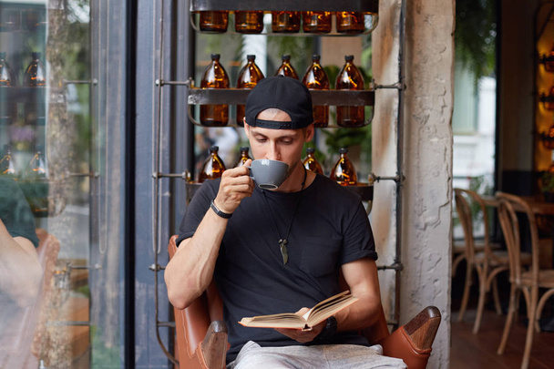 Расслабленный молодой бородатый кавказский парень наслаждается свободным временем, спокойной атмосферой на открытом воздухе, держит кружку с кофе, читает интересный роман, внимательно смотрит на книгу, сидит кресло над стильным интерьером кафе
.  - Фото, изображение