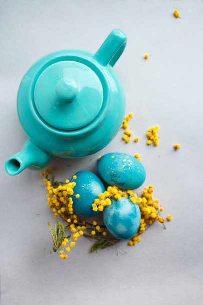 Easter composition: turquoise teapot and painted eggs on a gray background with mimosa flowers - Photo, image