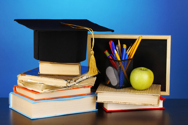 Books and magister cap against school board on wooden table on blue background - Foto, imagen