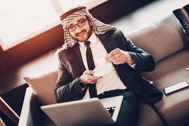 Businessman Using Laptop. Arab Businessman. Man Wearing in Black Suit. Experienced Entrepreneur. Successful Young Man. Work in Office .Arab in glasses. lying on couch. holding cup of coffee - 写真・画像