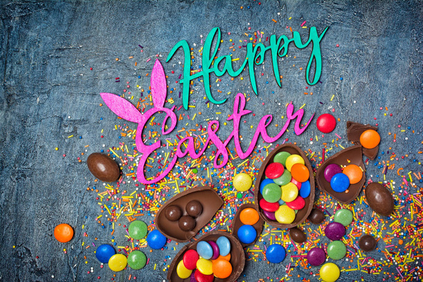 Top view on bright text of happy easter with chocolate traditional eggs and sprinkles for bakery on blue concrete background - Fotografie, Obrázek