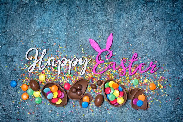 Top view on bright text of happy easter with chocolate traditional eggs and sprinkles for bakery on blue concrete background with copy space - Foto, afbeelding