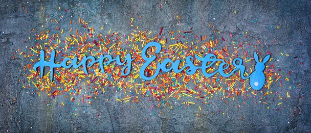Top view on bright text of happy easter with sugar sprinkles or confetti as decor for bakery on blue concrete background - Foto, Imagen
