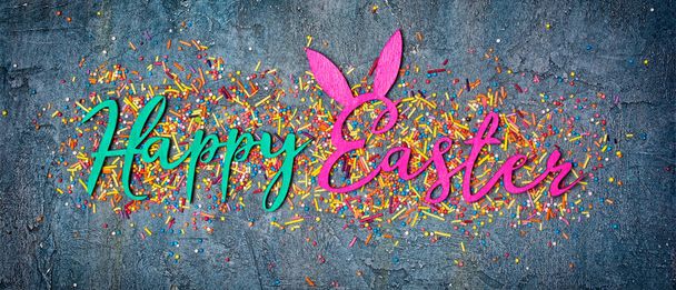 Top view on bright text of happy easter with sugar sprinkles or confetti as decor for bakery on blue concrete background - Foto, Bild