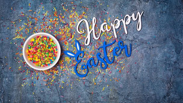 Top view on bright text of happy easter with sugar sprinkles or confetti in white bowl on blue concrete background with copy space - Fotografie, Obrázek