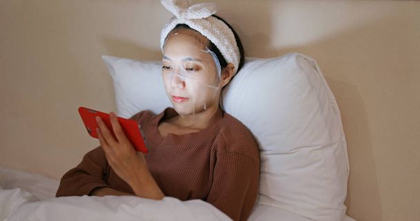 Woman uses mobile phone and applies face mask in bed at night - Φωτογραφία, εικόνα
