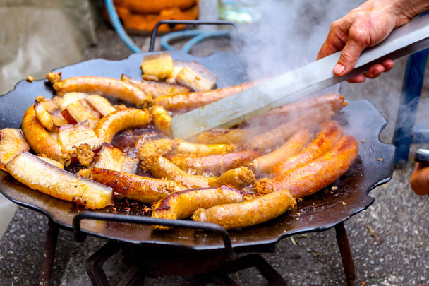 Hand with the help of metal tongs turns the simmering sausages on a black barbecue grill.  - Photo, Image