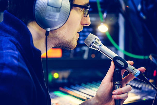Young man is sitting in the recording studio and talks into the microphone, buttons and equipment in the blurry background - Photo, image