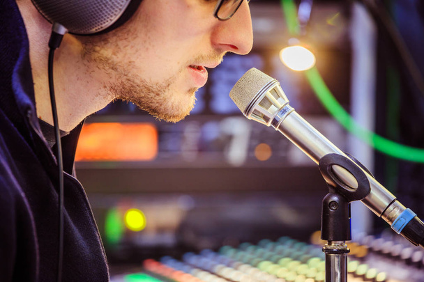 Young man is sitting in the recording studio and talks into the microphone, buttons and equipment in the blurry background - Valokuva, kuva
