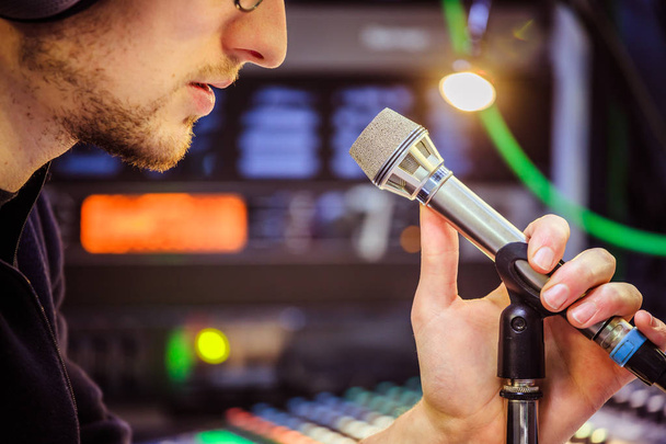 Young man is sitting in the recording studio and talks into the microphone, buttons and equipment in the blurry background - Photo, image