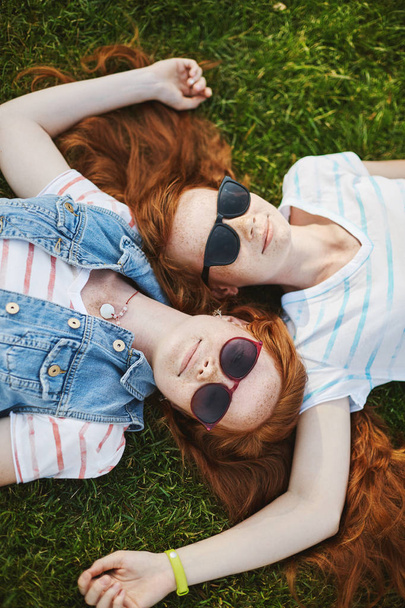 Vertical shot of beautiful relaxed and happy girls with natural ginger hair and cute freckles lying on grass, touching each other heads, wearing cool sunglasses, chilling under sun in warm summer day - Photo, image