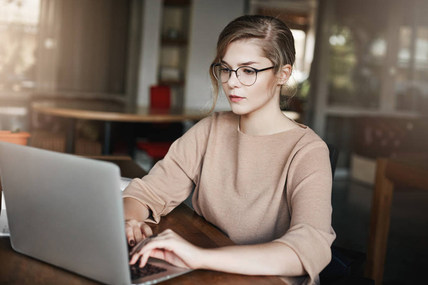 Дівчина знає, як керувати робочим часом. Студійний знімок привабливої серйозної європейської жінки в окулярах та офісному одязі, сидячи за столом, працюючи з ноутбуком та дивлячись орієнтованим на екран ноутбука
 - Фото, зображення