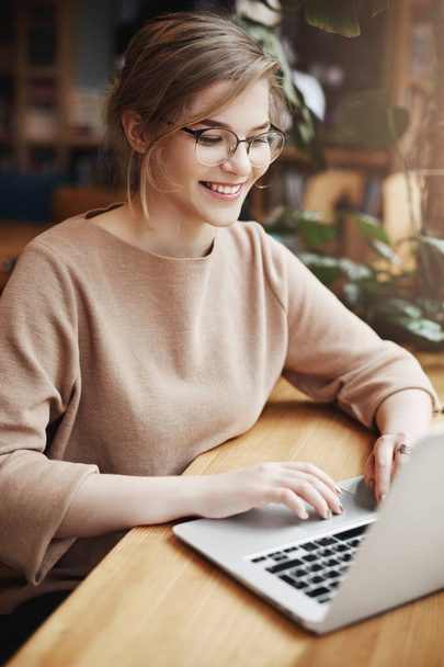 Vertical shot of successful and happy european female student in trendy glasses and casual outfit, smiling broadly while sitting near window in cafe and using laptop to update social page - Photo, image