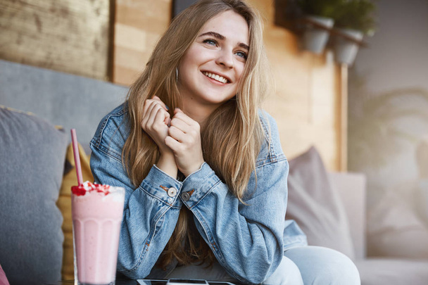 Woman easily falling in love, seeing charming man walking down street. Portrait of tender touched attractive dreamy girl with fair hair in denim jacket, sitting in cafe and drinking milkshake, smiling - Foto, imagen