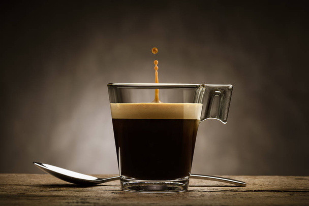Caffè nero in tazza di vetro con cucchiaino e goccia saltante, su tavolo di legno - Foto, immagini